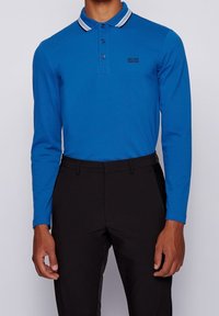 Långärmad pikétröja i klarblå bomull med randig krage, tvåknapps knäppning och en liten svart logotyp på vänster bröst.