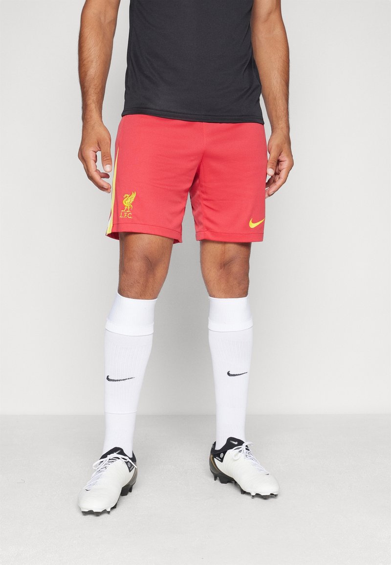 Mężczyzna w czerwonych szortach piłkarskich Liverpool FC, białych wysokich do kolan skarpetkach Nike, białych korkach piłkarskich oraz czarnej koszulce z krótkim rękawem na białym tle.