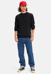 Black knitted sweater, blue jeans, and black sneakers. Red cap with white text. Casual fit, soft texture, and ribbed cuffs and hem.