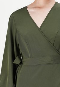 Olive green wrap dress made of smooth fabric, featuring long sleeves, a deep V-neck, and a tie belt at the waist.
