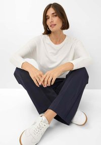 Haut à manches longues blanc, pantalon marine et baskets blanches, assis jambes croisées. Textures lisses, design minimal et couleurs neutres.