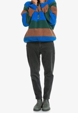 Persoon die een fleece trui met blauwe, groene en bruine strepen draagt, zwarte broek en grijze enkellaarzen, staande met handen bij de kraag.