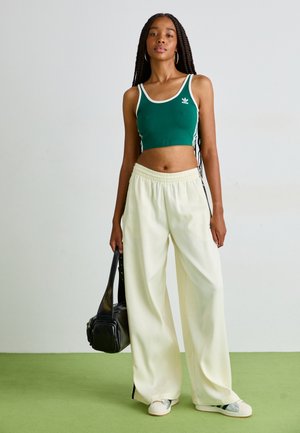 Green cropped tank top with white trim, paired with wide-leg cream trousers. Model holds a small black bag. Simple sneaker shoes.