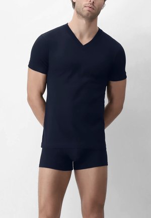 Uomo che indossa una maglietta aderente a maniche corte di colore blu navy scuro con scollo a V e boxer abbinati, in piedi con le mani dietro la schiena su sfondo chiaro.