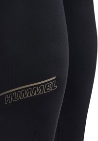 Mustade spordipõlvikute lähivaade, millel on reie piirkonnas kullas värvitud kaubamärgi nimi "HUMMEL".