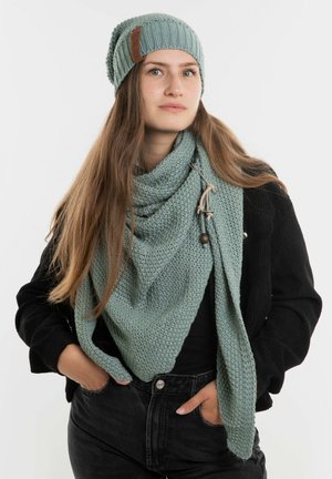 Gehaakte mintgroene sjaal om de nek van een model, met een gestructureerd patroon. Bijpassende beanie met een bruine leren patch accenten.