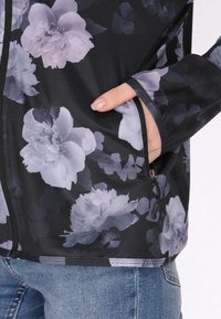 Giacca floreale in tessuto nero con motivi di fiori grigi e viola. Presenta una tasca con zip e polsini aderenti.