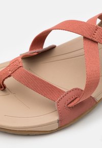 Koralli risti-nauha sandaali, jossa on teksturoitu pinta, pehmeä beige pohja ja kestävä kumipohja. Nauhoissa on ompelutaksennuksia.