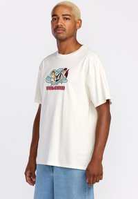 Camiseta de algodón crema con mangas cortas. Presenta un diseño gráfico de ángeles y demonios con el texto "VOLCOM" en rojo. Corte relajado.