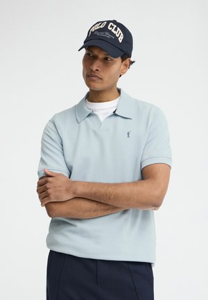 Hombre que lleva una camisa polo azul claro encima de una camiseta blanca y una gorra azul marino con el texto "Polo Club", brazos cruzados, mirando ligeramente hacia un lado.