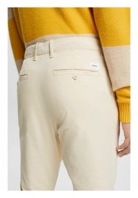 Pantaloni beige con due tasche posteriori e chiusura a bottone. Il tessuto è liscio con una leggera lucentezza. Indossati con un maglione a strisce giallo brillante.