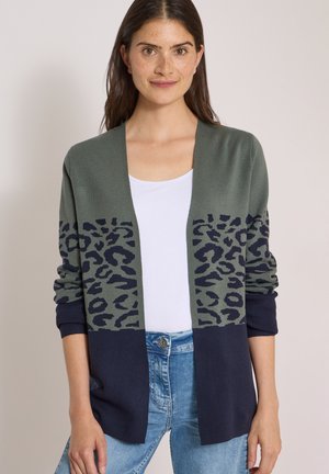 Donna con cardigan aperto davanti con stampa leopardata verde e blu navy, top bianco e jeans azzurri, in piedi davanti a uno sfondo semplice.
