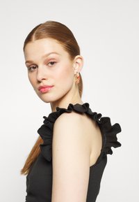 Top de tanque preto com textura canelada e detalhes de ombreira franzida. Modelo usa brincos coloridos e translúcidos. Cabelo liso preso em um rabo de cavalo.