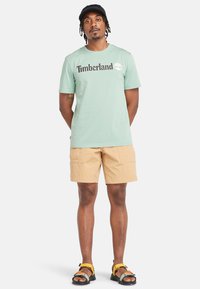 Mátově zelené bavlněné tričko s logem "Timberland", béžové cargo šortky a černé sandály s žlutými a modrými akcenty.