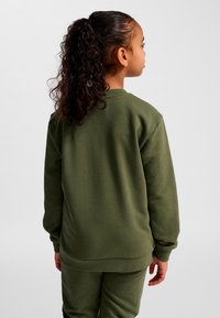 Ung flicka med lockigt hår uppsatt, iklädd olivgrön sweatshirt och matchande byxor, sedd bakifrån mot en enfärgad bakgrund.