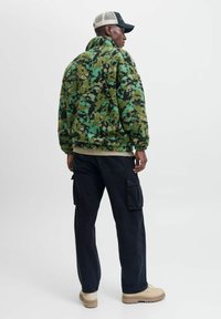 Fleece jack in groen camouflagepatroon, hoge kraag, geribbelde mouwen, gecombineerd met donkere cargo pants en beige laarzen. Achteraanzicht.