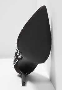 Chaussure à talon haut noire à bout pointu avec des clous ronds argentés décorant la partie supérieure, vue de la semelle face à l'appareil photo.