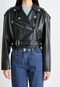 Giacca da motociclista in similpelle nera con dettagli argentati, collo con revers, zip laterali e sul petto, vita con cintura, abbinata a jeans blu.