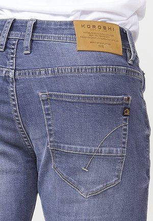 Jeans da uomo in denim blu con un'etichetta in pelle che riporta la scritta "KOROSHI". Presentano una tasca posteriore con dettagli di cucitura e un piccolo accento di etichetta.