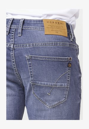 Jeans de mezclilla azules para hombre con una etiqueta de cuero que dice "KOROSHI." Presenta un bolsillo trasero con detalles de costura y un pequeño acento de etiqueta.