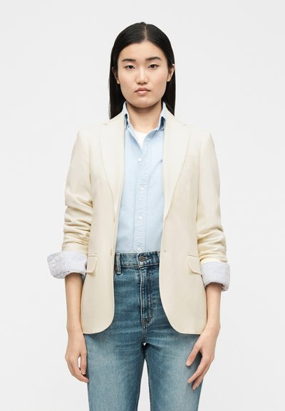Νεαρή γυναίκα που φοράει ένα κρεμ σακάκι πάνω από ένα ανοιχτό γαλάζιο πουκάμισο με κουμπιά και ψηλόμεσα μπλε τζιν, στέκεται μπροστά σε απλό λευκό φόντο.