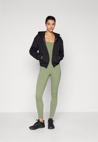Black zip-up hoodie with front pockets, worn over a fitted olive green bodysuit and matching leggings, paired with black athletic shoes.