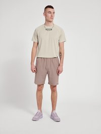 Beige träningströja med korta raglanärmar och diskret logotyp, ihop med ljusbruna shorts. Skorna är ljuslila sneakers.