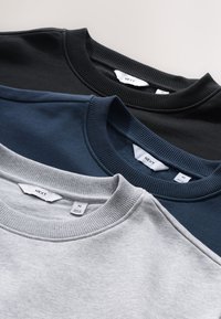 Tre felpe girocollo in nero, blu navy e grigio chiaro, con polsini e collo a costine, con etichette in tessuto visibili sui colletti.