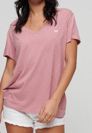 Mujer con una camiseta rosa de cuello en V holgada con el logo "Super Dry" y shorts de mezclilla blancos con bordes deshilachados, posando frente a un fondo gris.