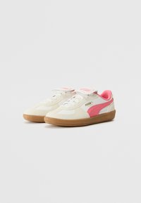 Baskets blanches avec un accent rose et un logo doré, dotées d'une semelle en caoutchouc texturé, de lacets et d'une sangle en velcro pour un maintien sécurisé.