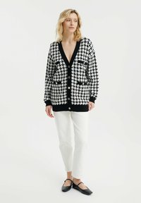 Crni i bijeli kardigan s uzorkom houndstooth, dugih rukava, s dva prednja džepa i crnim gumbima. Uparen s bijelim hlačama i crnim balerinama.