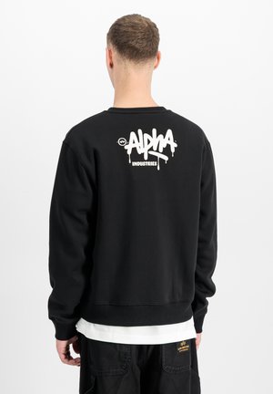 Man wearing black sweatshirt with white "Alpha Industries" logo on the upper back, standing against a plain white background.