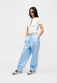 Γυναίκα με μακριά μαύρα μαλλιά, φοράει λευκό κοντό t-shirt, ανοιχτό γαλάζιο παντελόνι Jordan, αθλητικά παπούτσια και σκούρα γυαλιά ηλίου, στέκεται με τα χέρια στις τσέπες.