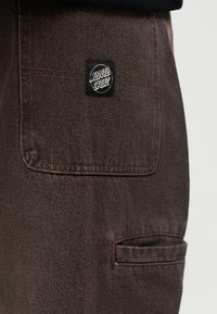 Barna farmeranyag egy fekete Santa Cruz folttal, amelyet egy ruhadarab felső zsebére varrtak.