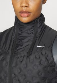 Chaleco acolchado negro con cuello alto, cierre de cremallera y patrón de puntos en relieve. Presenta un logo blanco de Nike en el pecho. Textura elegante y suave.