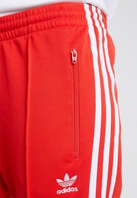 Röda sweatpants gjorda av tyg med en slät textur. Har vita ränder och en ficka med dragkedja på sidan. Adidas-logotypen vid underbenet.