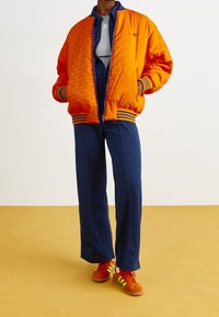 Orange quilted bomber jacket with navy stripes at cuffs, paired with navy wide-leg pants and orange sneakers with green accents.
