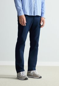 Pantaloni bleumarin dintr-un material neted, asortați cu adidași gri și albi. Cămășa este de culoare albastră deschisă, cu dungi verticale.