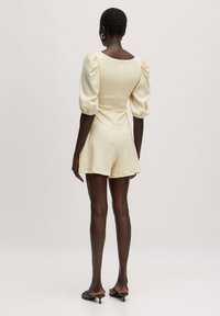 Crèmekleurige playsuit met puffmouwen, een aansluitend lijfje en korte shortjes. Achterzak met ritssluiting en gemêleerde stof benadrukken het ontwerp.