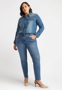 Camisa de mezclilla abotonada con dos bolsillos en el pecho y acentos dorados, combinada con jeans azules que presentan detalles de cadena en los bolsillos y tacones nude puntiagudos.