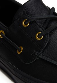 Gros plan d'une chaussure en cuir noir avec des lacets noirs enfilés à travers des œillets métalliques dorés.