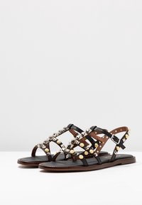 Sandalias negras con tiras y suela plana, que presentan tachuelas metálicas y acentos dorados. Las tiras están hechas de cuero con un diseño cruzado.