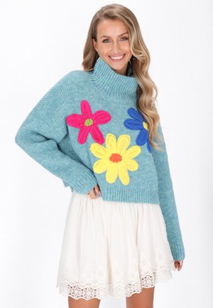 Maglione lavorato a maglia turchese con collo alto e vestibilità oversize, decorato con applicazioni floreali colorate in rosa, blu e giallo. Gonna di pizzo bianca.