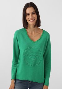 Grüner Pullover-Sweatshirt mit V-Ausschnitt, mit einem geprägten Textdesign in der Mitte der Brust und ausgefransten Rändern entlang des Halsausschnitts und Saums.