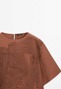 Haut en lin marron présentant des plis tourbillonnants texturés sur le devant, avec un col rond et des manches courtes. Fermeture zippée au dos.