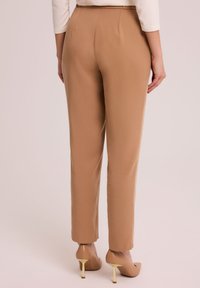 Beżowe spodnie o dopasowanym kroju i prostych nogawkach, z wyraźnym zagięciem z przodu, gładką fakturą i średnim stanem. Noszone z cielistymi szpilkami.