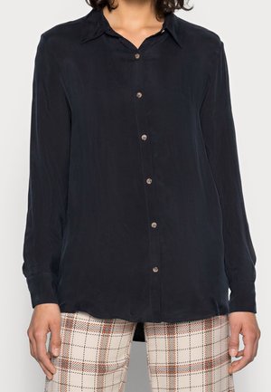 Donna che indossa una camicia blu navy a maniche lunghe con bottoni abbinata a pantaloni beige a quadri, in piedi davanti a uno sfondo semplice.