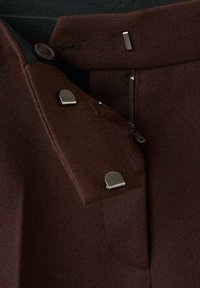 Smeđi tkaninski hlače s bočnim patentnim zatvaračem, s metalnim gumbima i detaljima. Materijal ima teksturiranu površinu i kroj prilagođen tijelu.
