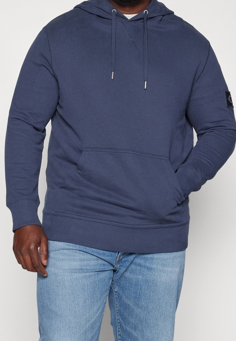 Sudadera con capucha azul marino hecha de tela suave, con un bolsillo canguro, puños acanalados y un pequeño parche con logo en la manga.