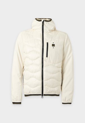 Jeune homme debout, mains dans les poches, portant une veste matelassée crème, une chemise blanche, un jean bleu clair ample et des baskets blanches.